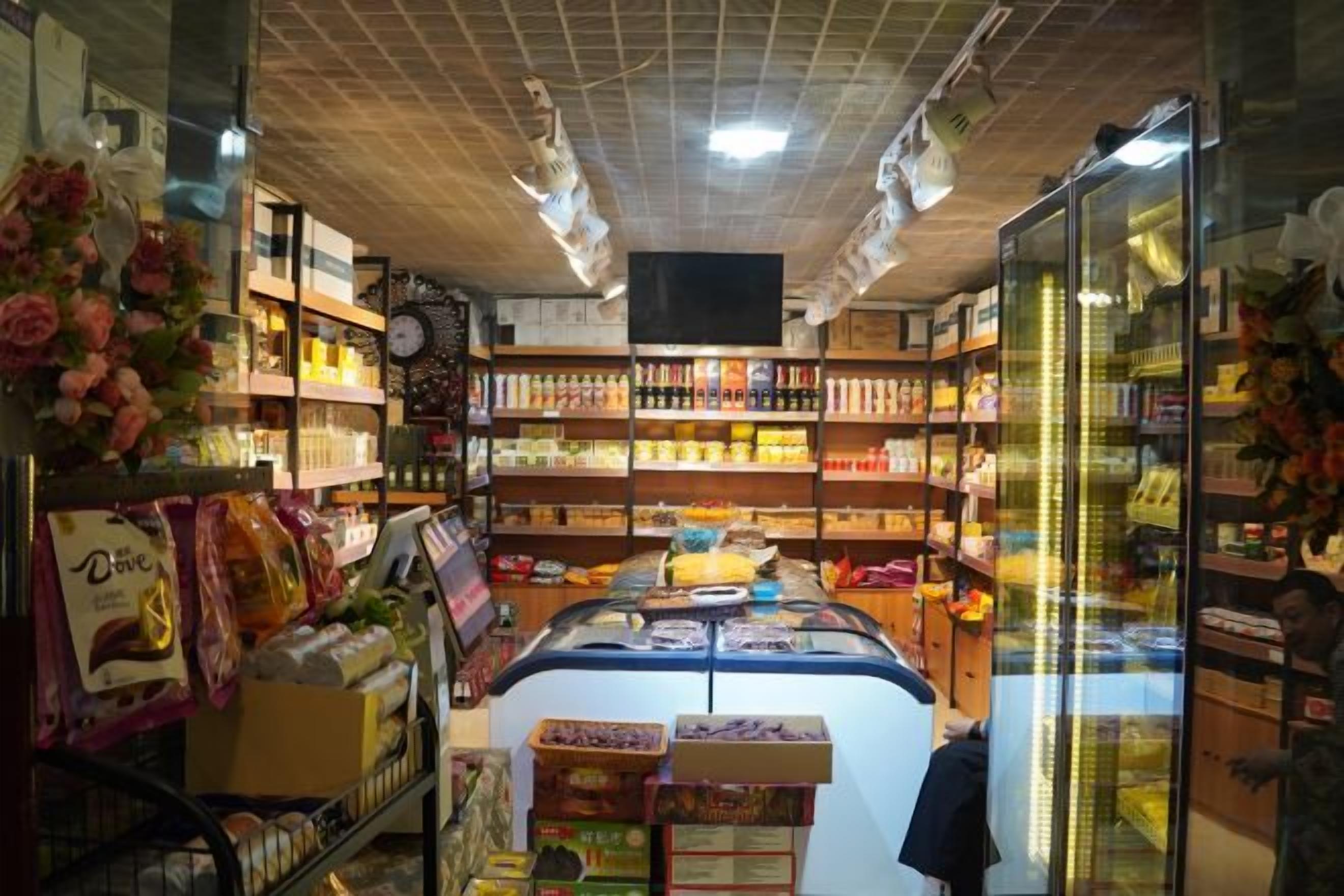 A grocery store with shelves stocked with various products, a refrigerated display case, and a checkout area.