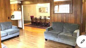 A living room with two grey sofas, a wooden floor, a dining area with a table and chairs, and a large window.