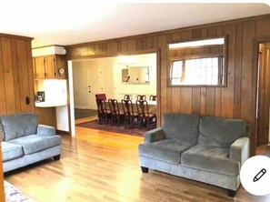 A living room with two grey sofas, a wooden floor, a dining area with a table and chairs, and a large window.