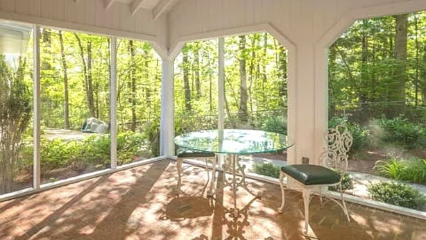 A covered patio with a glass table and chairs, surrounded by large windows offering a view of a forested area.