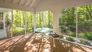 A covered patio with a glass table and chairs, surrounded by large windows offering a view of a wooded area.