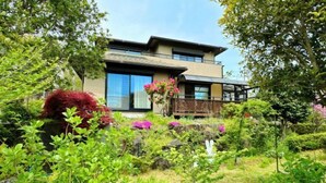 Une maison moderne à deux étages, dotée d’un balcon et entourée d’une végétation luxuriante et de plantes en fleurs.