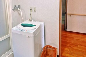 A compact, white washing machine in a corner with a wooden floor and a basket nearby. - Vacation Rental Hanatsusuri Izukogen (Ito)