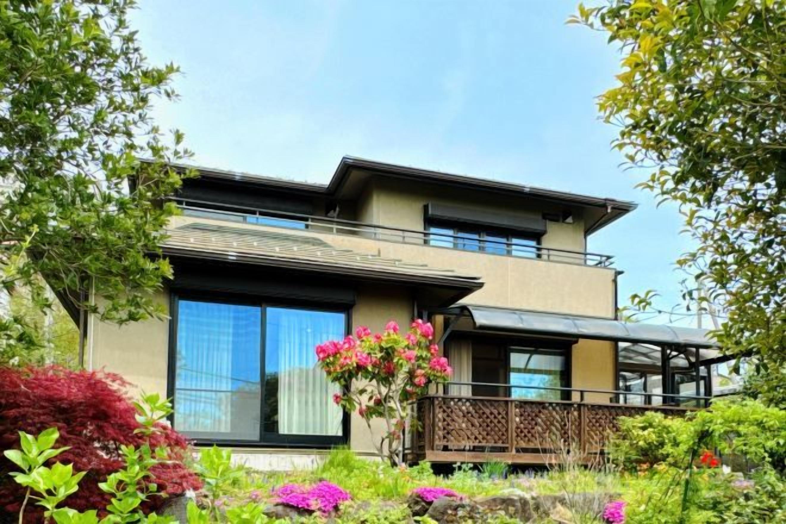 A modern two-story house with a balcony, surrounded by lush greenery and flowering plants.