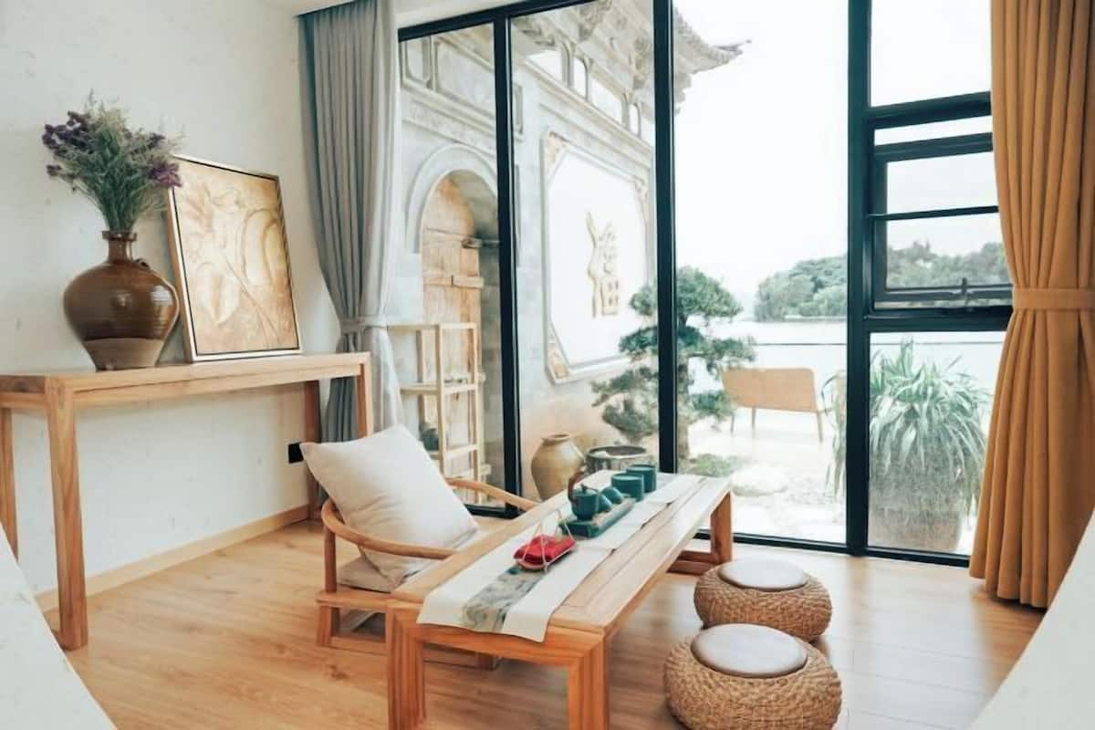 A room with a wooden bench, a vase with flowers, a framed picture, and a view of a building through large windows.