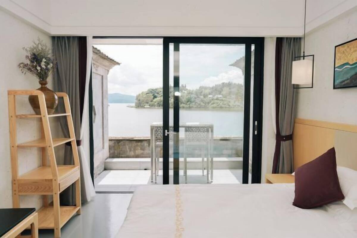 A hotel room with a bed, a wooden ladder, a table with chairs, and a view of a lake through a glass door.