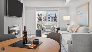 A modern hotel room with a flat-screen TV, a fireplace, a dining table with a bottle of wine and glasses, a sofa, and a large window with a view of buildings. - 1 Bed/1 Bath Sparrowhawk-Courtyard Facing-Sleeps 4 (Dead Man's Flats)