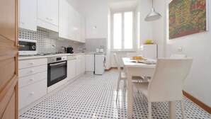 Cocina compacta con gabinetes blancos, microondas, estufa y pileta. Cuenta con un comedor con mesa y sillas, y un ventanal que proporciona luz natural.