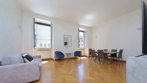Amplia sala de estar con sofá, mesa de comedor y sillas. Grandes ventanales permiten la entrada de luz natural.