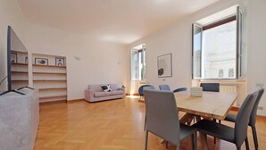 Amplia sala de estar con piso de madera, un sofá, una mesa de comedor con sillas y un ventanal con vista a un edificio.