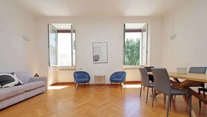 Amplia sala de estar con piso de madera, un sofá, dos sillas azules, una mesa de comedor con sillas y ventanas grandes que ofrecen vista a árboles.