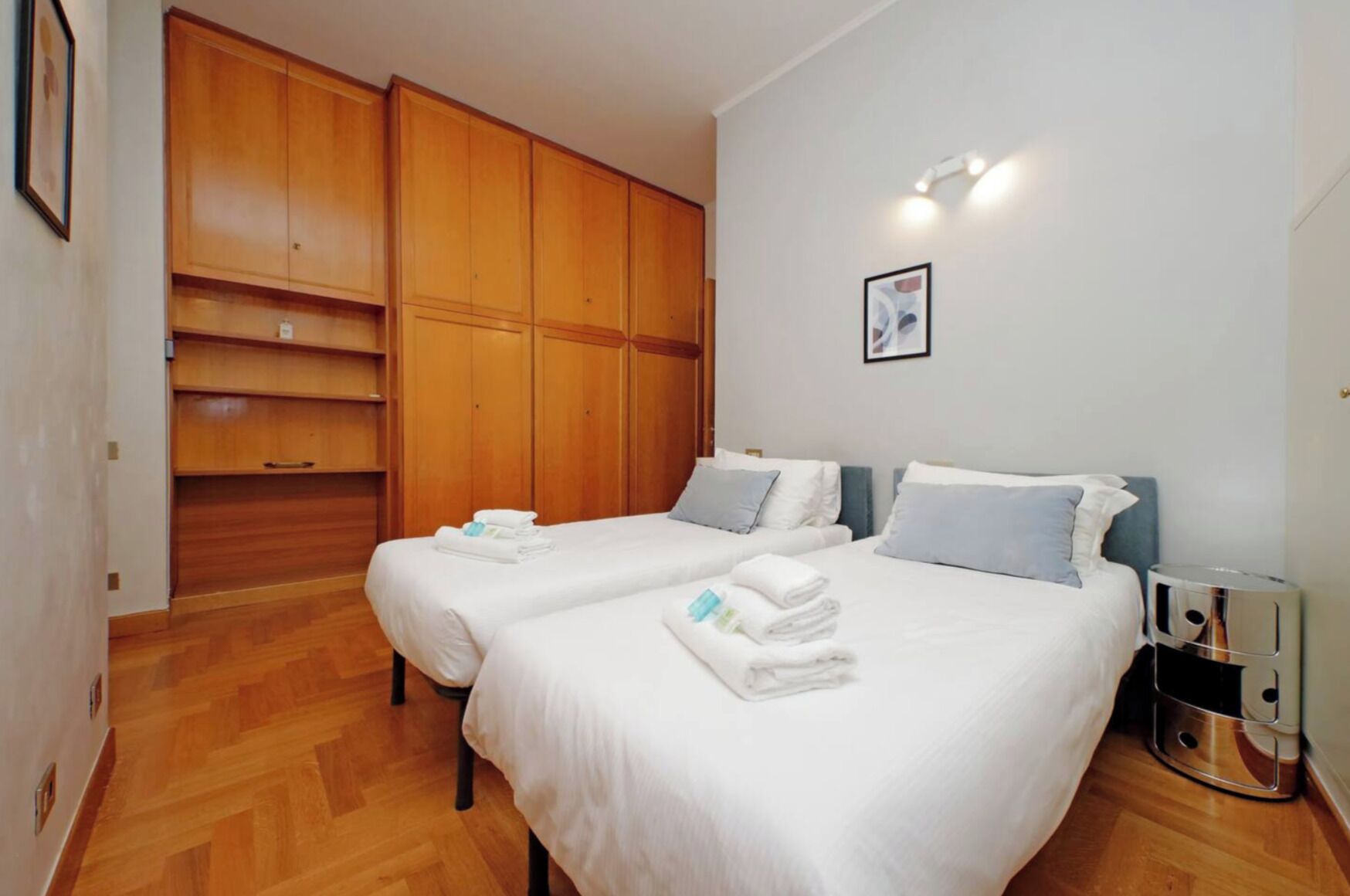 A hotel room with two beds, wooden wardrobe, and a bedside table with a lamp.