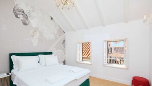 A bedroom with a green bed frame, white bedding, two windows with a view of a courtyard, and a red chair. - Karma Suites by Dominium Comfort double room (Dubrovnik)