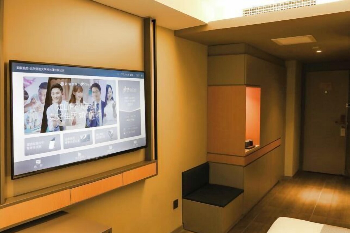Ein Hotelzimmer mit einem großen Flachbildfernseher an der Wand, einer kleinen Sitzecke mit gepolstertem Sitz und einer Holzkabine mit Lampe.