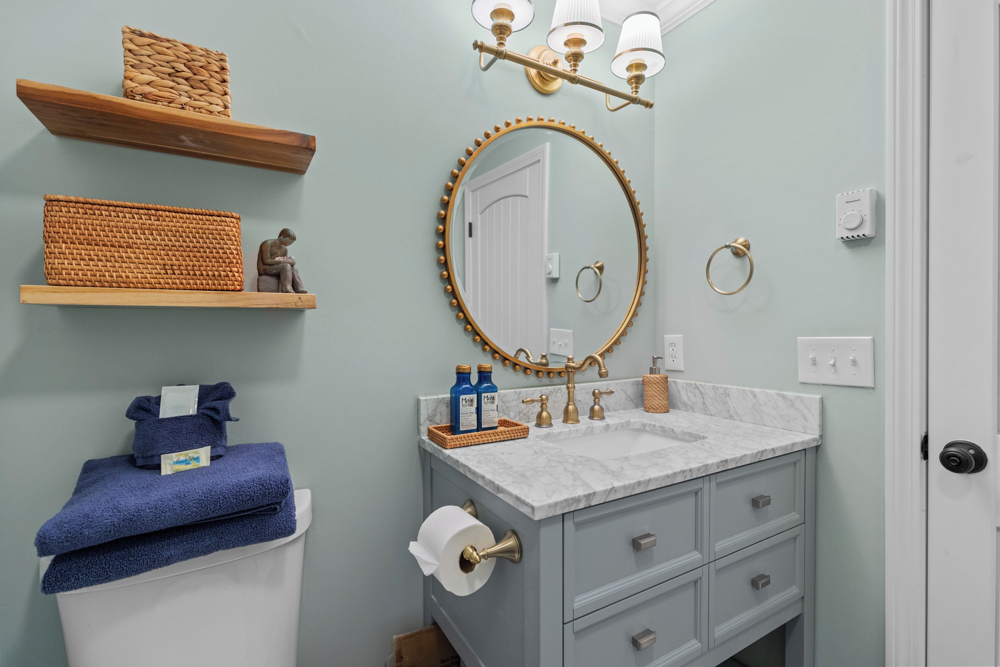 A bathroom with a marble sink, a round mirror with a gold frame, and wooden shelves with baskets.