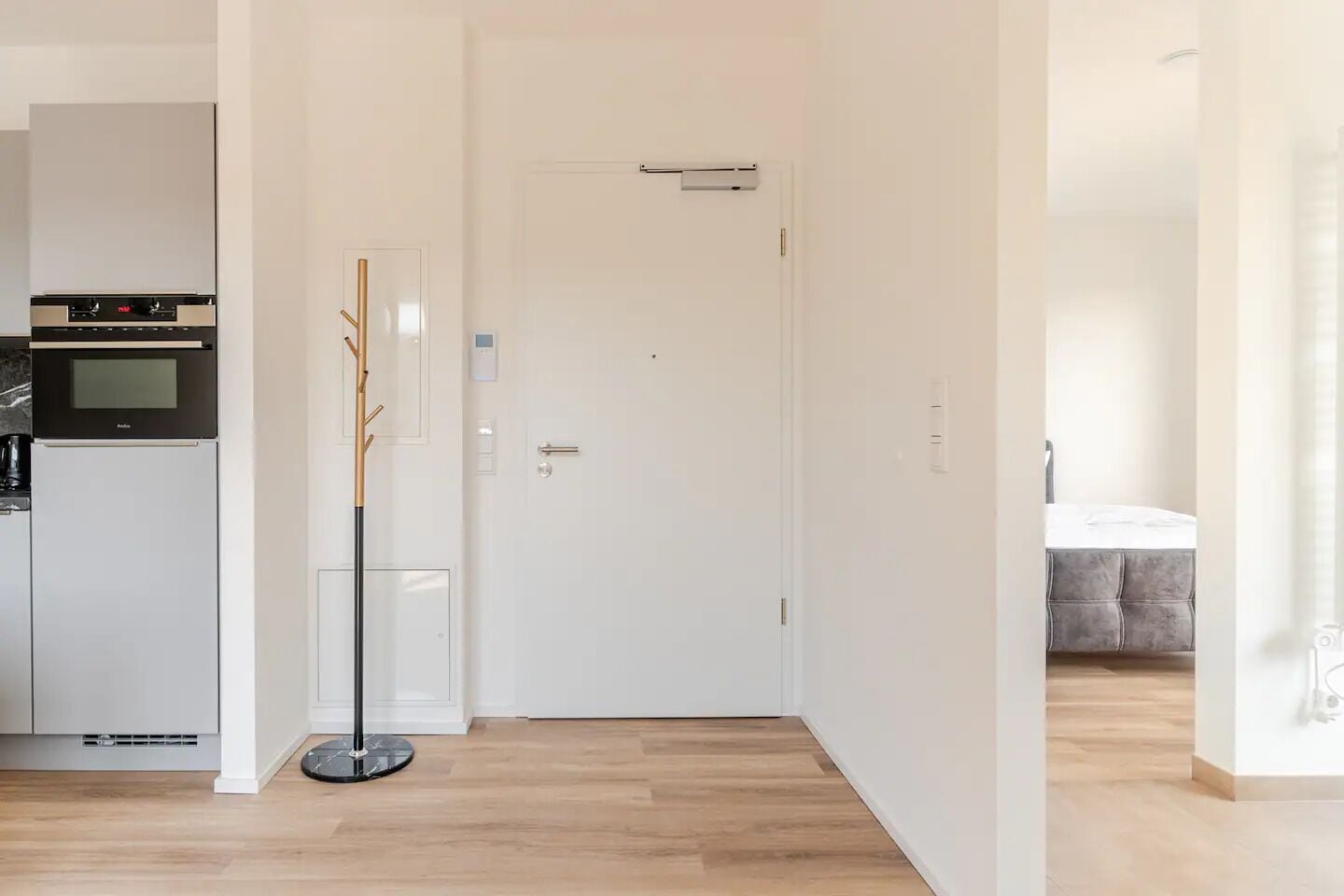 Una cocina moderna con horno empotrado y microondas, una puerta blanca, y un pasillo que conduce a un dormitorio con cama y mesita de noche.