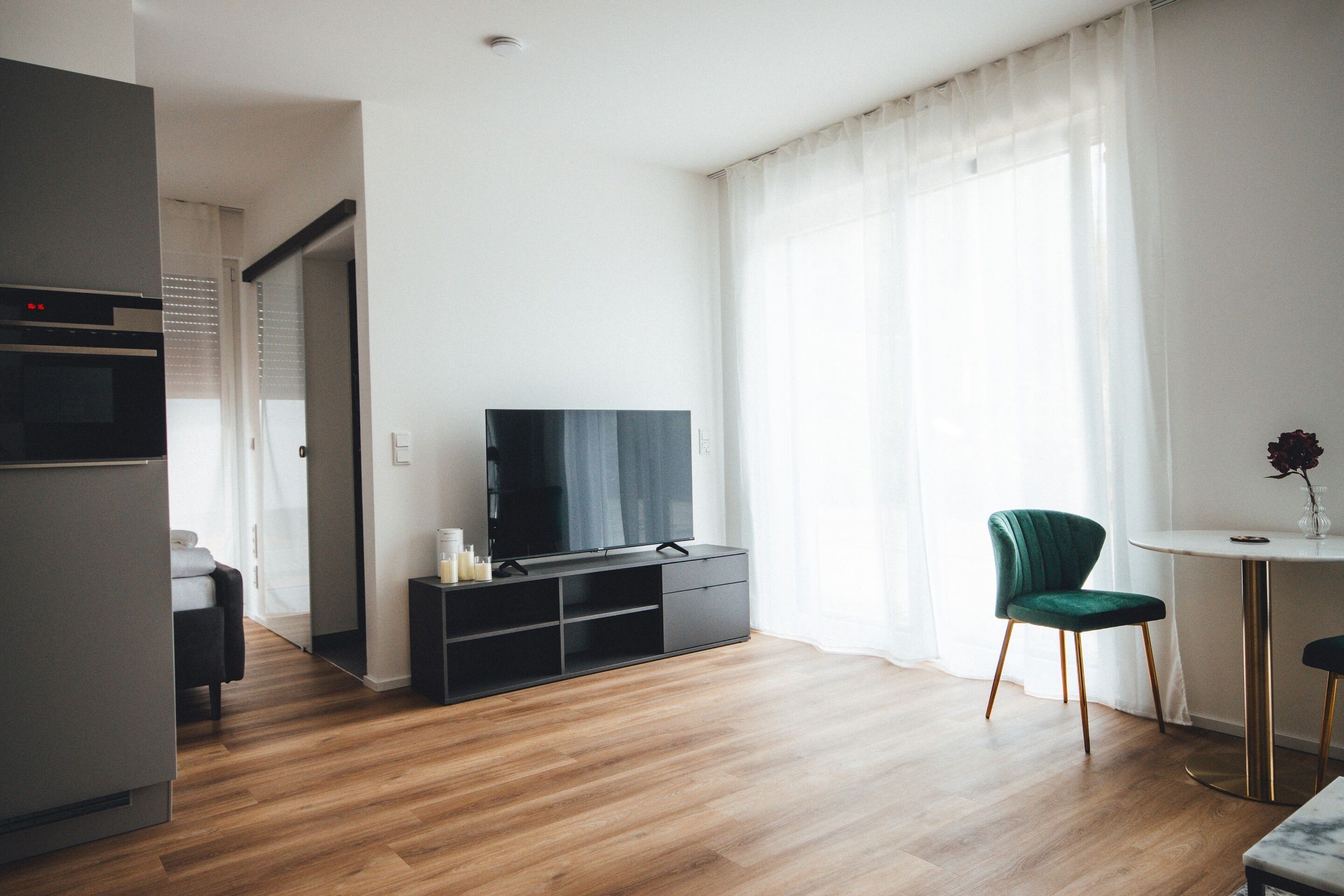 Ein modernes Wohnzimmer mit einem Flachbildfernseher auf einer schwarzen Unterhaltungsecke, einem grünen gepolsterten Sessel, einem kleinen runden Tisch mit einer Vase und Holzböden.