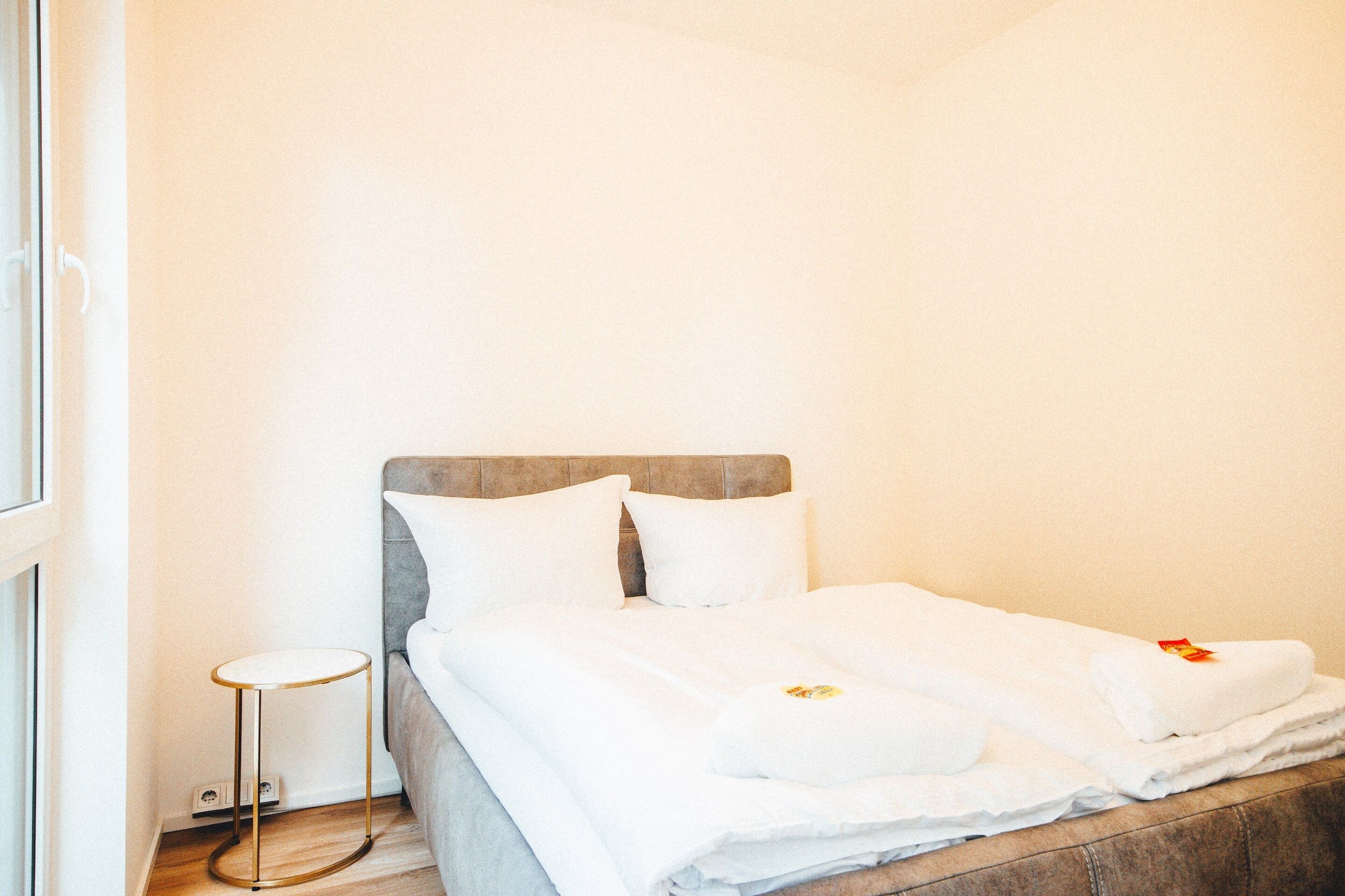 Un lit bien fait, avec du linge de lit blanc et des oreillers, une petite table d’appoint ronde et une tête de lit en bois.