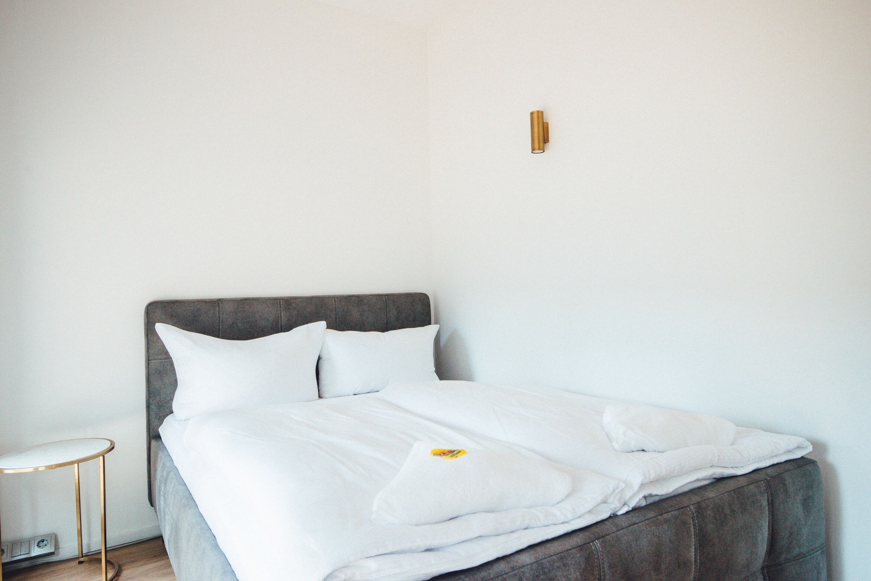 A neatly made bed with white bedding and pillows, a small bedside table with a lamp, and a wall-mounted light fixture.