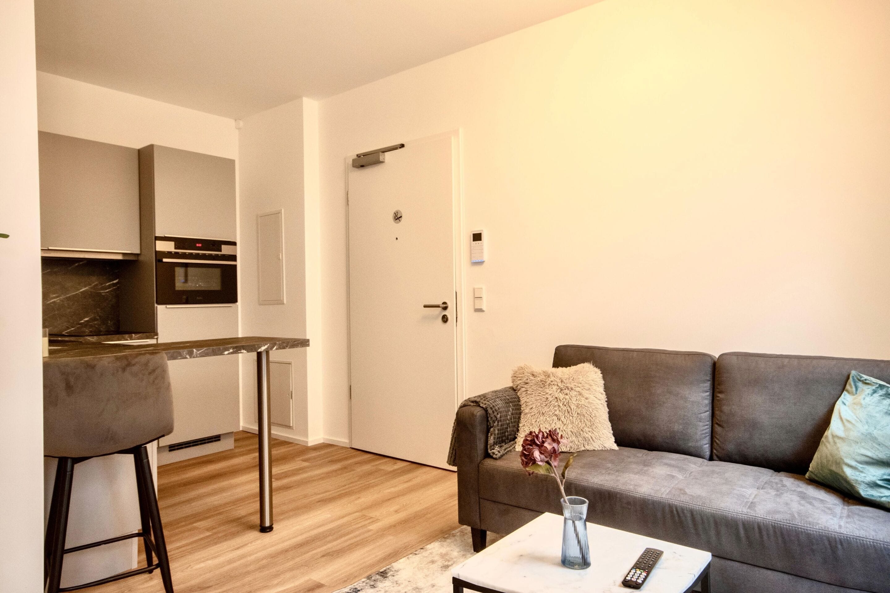 Una sala de estar moderna con un sofá gris, una pequeña mesa de centro y una cocina al fondo.