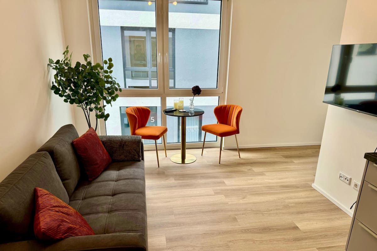 Un salon moderne avec un canapé marron foncé, deux fauteuils orange, une petite table ronde et une grande fenêtre donnant sur un immeuble.