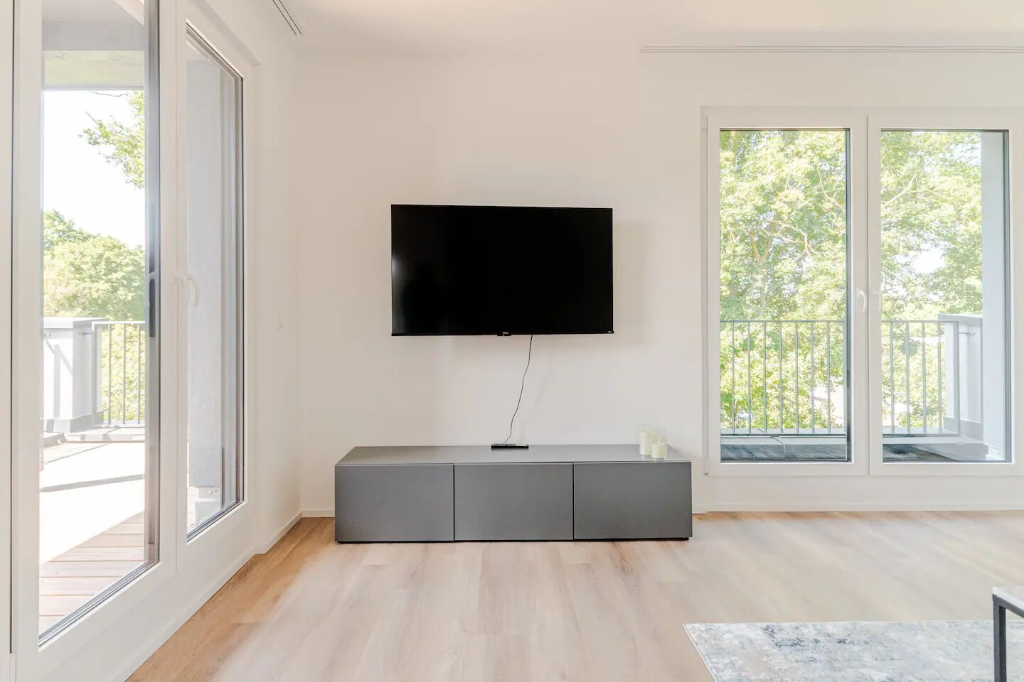 Un soggiorno moderno con una televisione a schermo piatto montata a parete, un mobile TV grigio e ampie finestre che offrono vista sull'esterno.