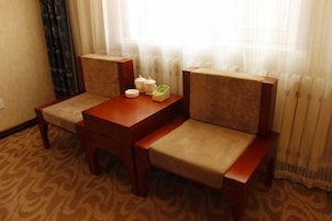Habitación de hotel con una mesa de madera y dos sillones tapizados, una mesita de noche con algunos objetos, y una ventana con cortinas translúcidas.