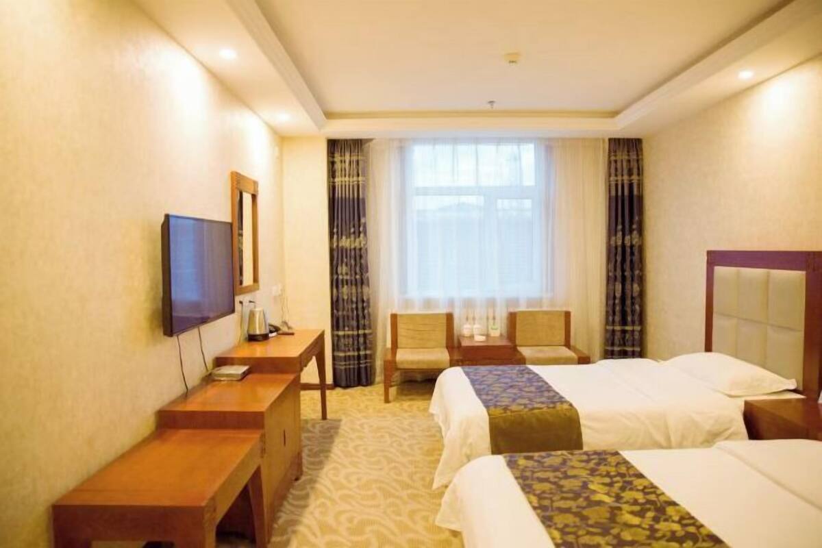 A hotel room with two beds, a desk, a television, and a large window with curtains.