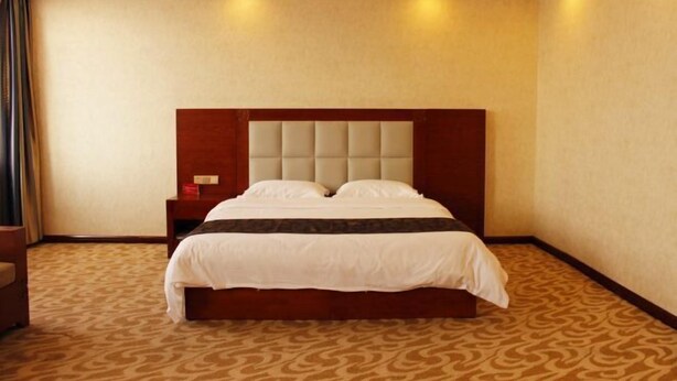A hotel room with a large bed, a wooden headboard, a nightstand, and a patterned carpet. - Yake Shiyuxin Business Hotel (Yakeshi)