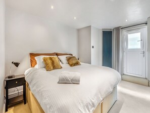 A neatly made bed with white linens and two decorative pillows, a bedside table with a lamp, and a view of a door with curtains. - 1 bedroom accommodation in Bowness-on-Windermere (Bowness-on-Windermere)