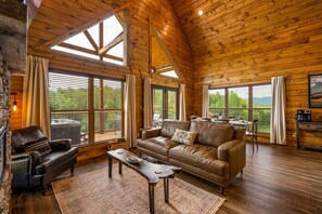 L’intérieur chaleureux d’un chalet avec un canapé en cuir, une cheminée en pierre et de grandes fenêtres offrant une vue sur l’extérieur.