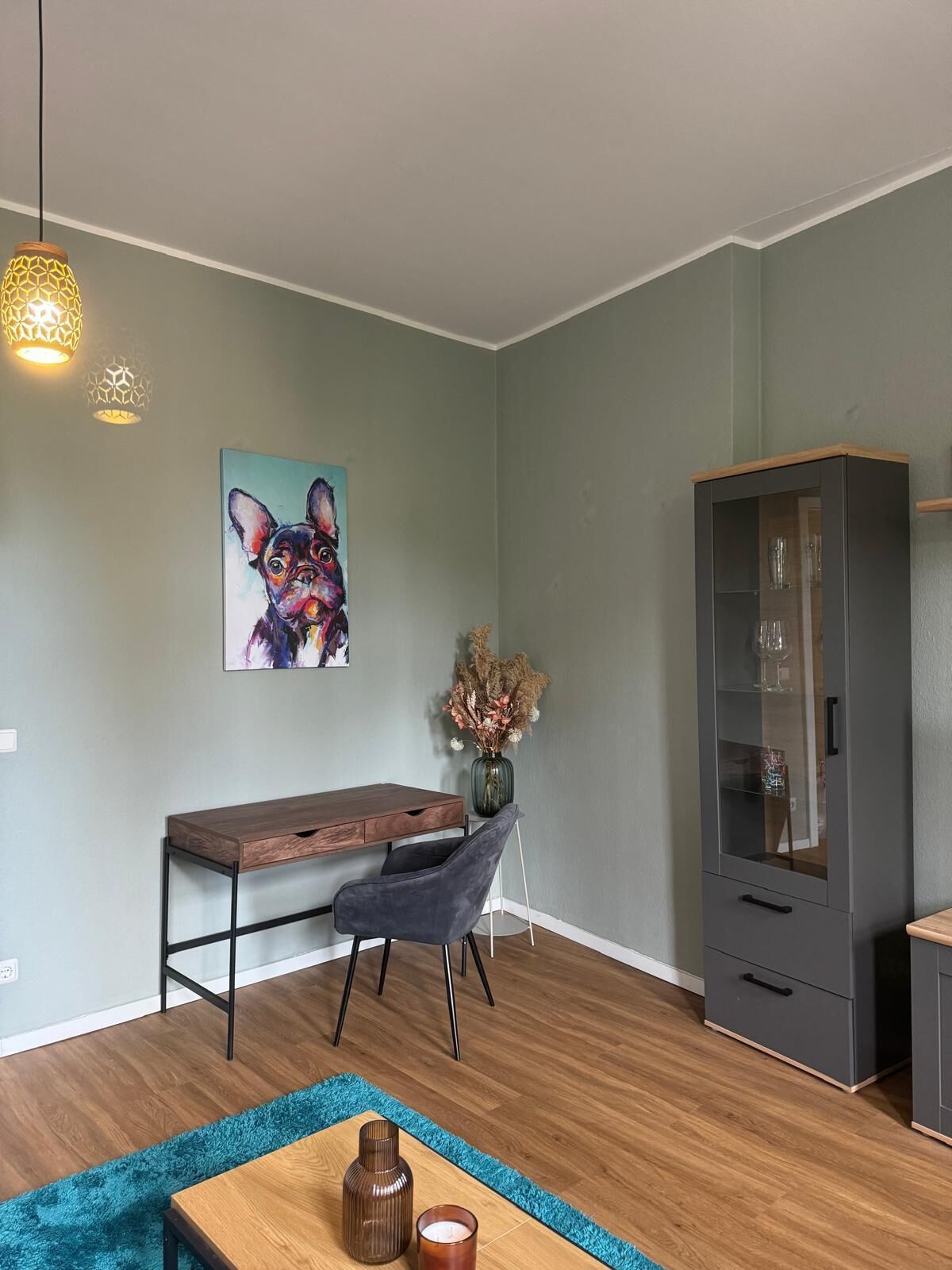 A modern living room with a wooden desk, a grey armchair, a cabinet with glass doors, and a painting of a dog on the wall.