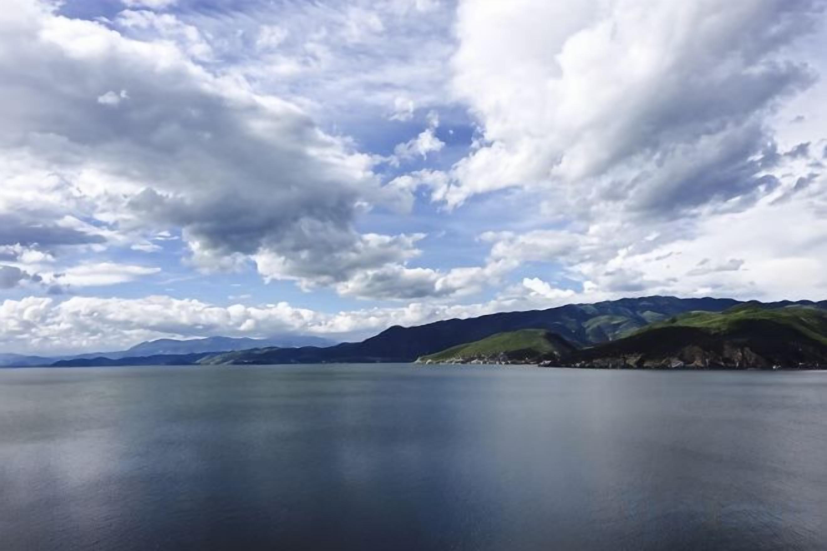 A calm lake with a mountain range in the background and a partly cloudy sky.