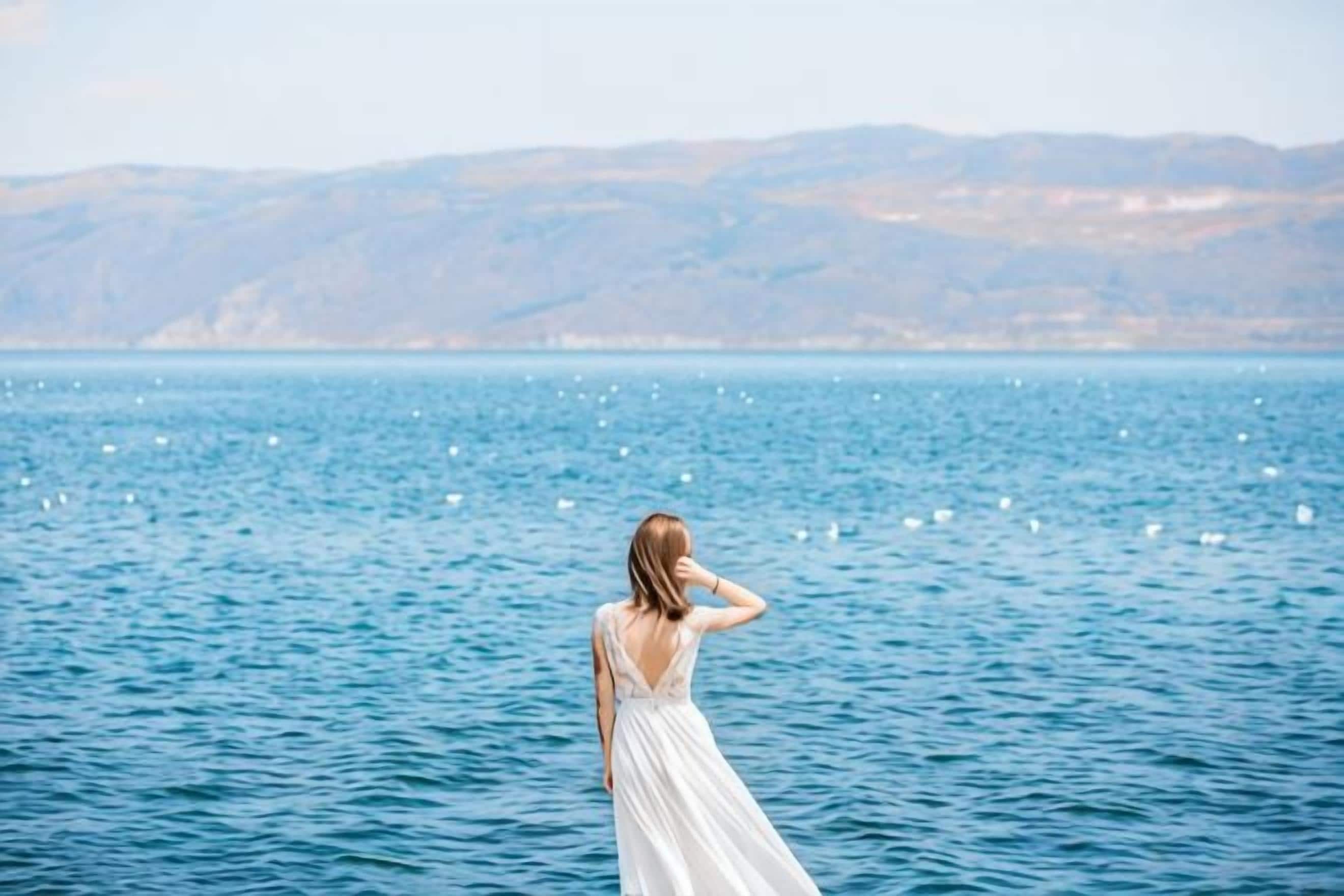 Una persona amb un vestit blanc dret al costat d'una massa d'aigua, amb muntanyes al fons.