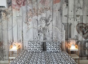 Une chambre à coucher avec un lit doté d’une tête de lit en métal blanche ornée d’un motif en forme de cœur, flanqué de deux tables de chevet avec des lampes. Les murs sont décorés de motifs floraux et en forme de cœur.