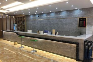 Een hotel receptie met een marmeren balie, een digitaal scherm en een zithoek met krukjes.