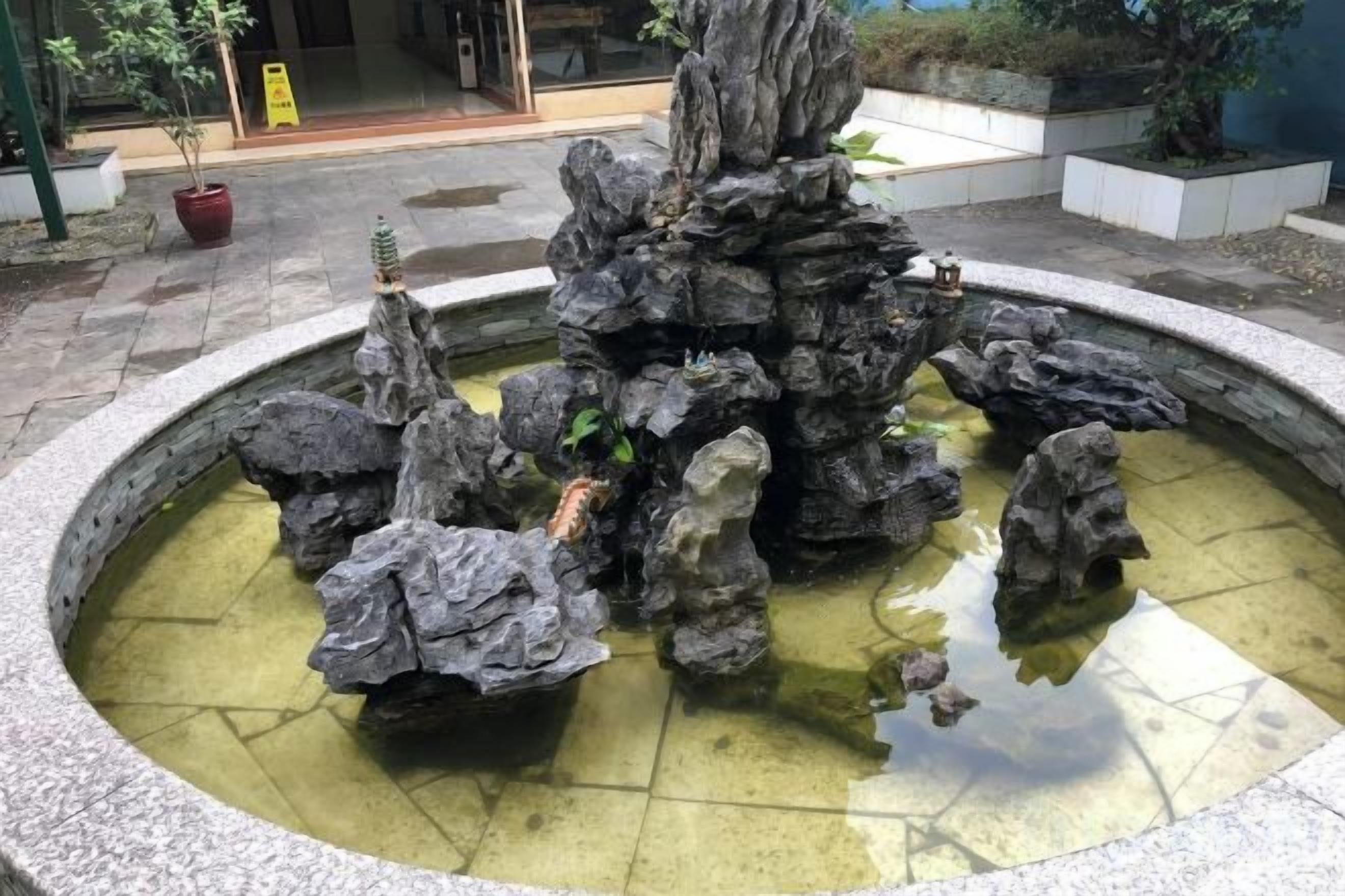 Une fontaine circulaire en pierre, avec une formation rocheuse centrale et des rochers disposés tout autour, le tout étant entouré d’une zone pavée.