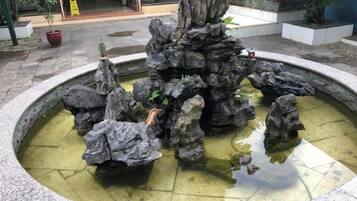 Une fontaine circulaire en pierre, avec une formation rocheuse centrale et des rochers disposés tout autour, le tout étant entouré d’une zone pavée.