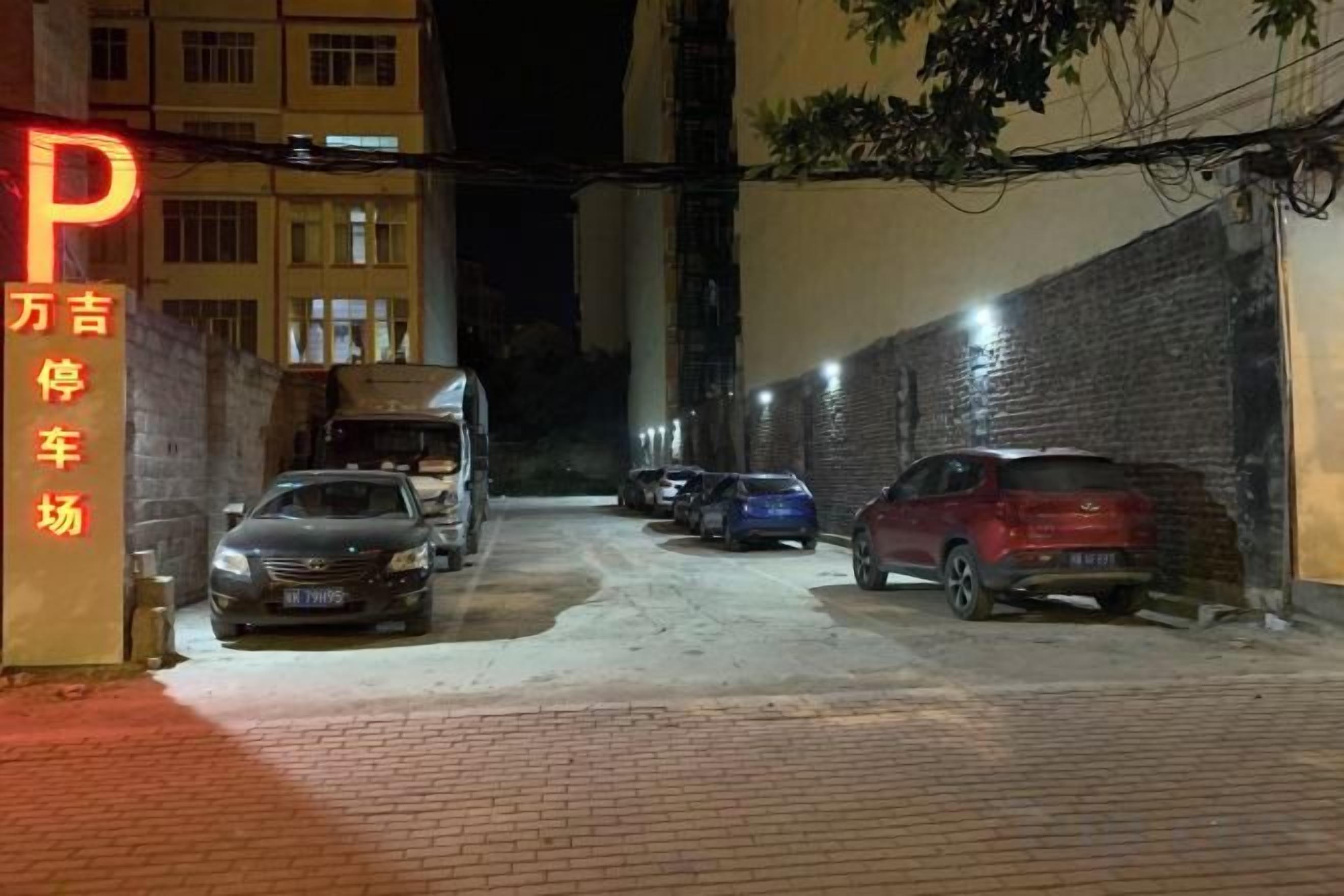 Un área de estacionamiento con varios autos y un camión, un edificio con un letrero con la letra 'P', y un muro de ladrillo.