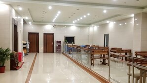 Un vestíbulo amplio con piso pulido, bancos de madera, barandilla de vidrio y una zona de recepción con mostrador y plantas en macetas.