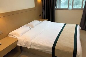 Un lit bien fait, avec une literie blanche et une bande verte, une table de chevet avec une lampe, et une fenêtre avec des rideaux.