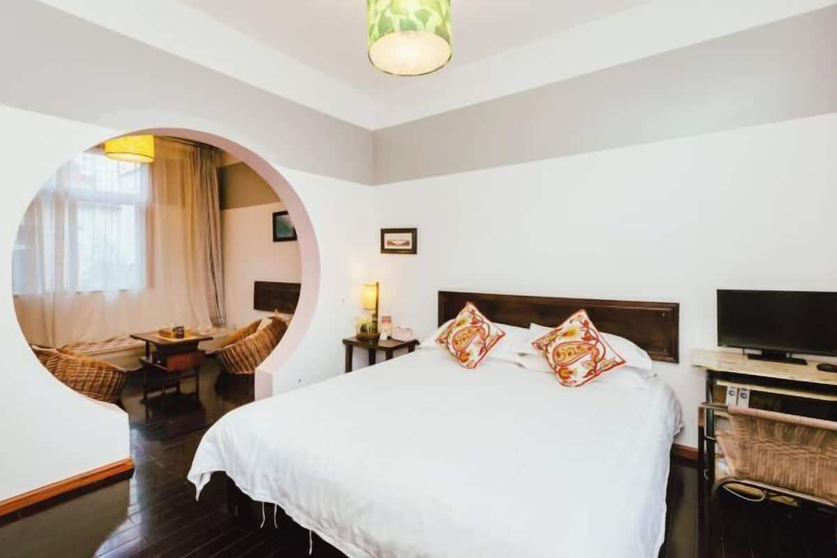 A hotel room with a large bed, a television, a desk with a chair, and a circular window overlooking a balcony.