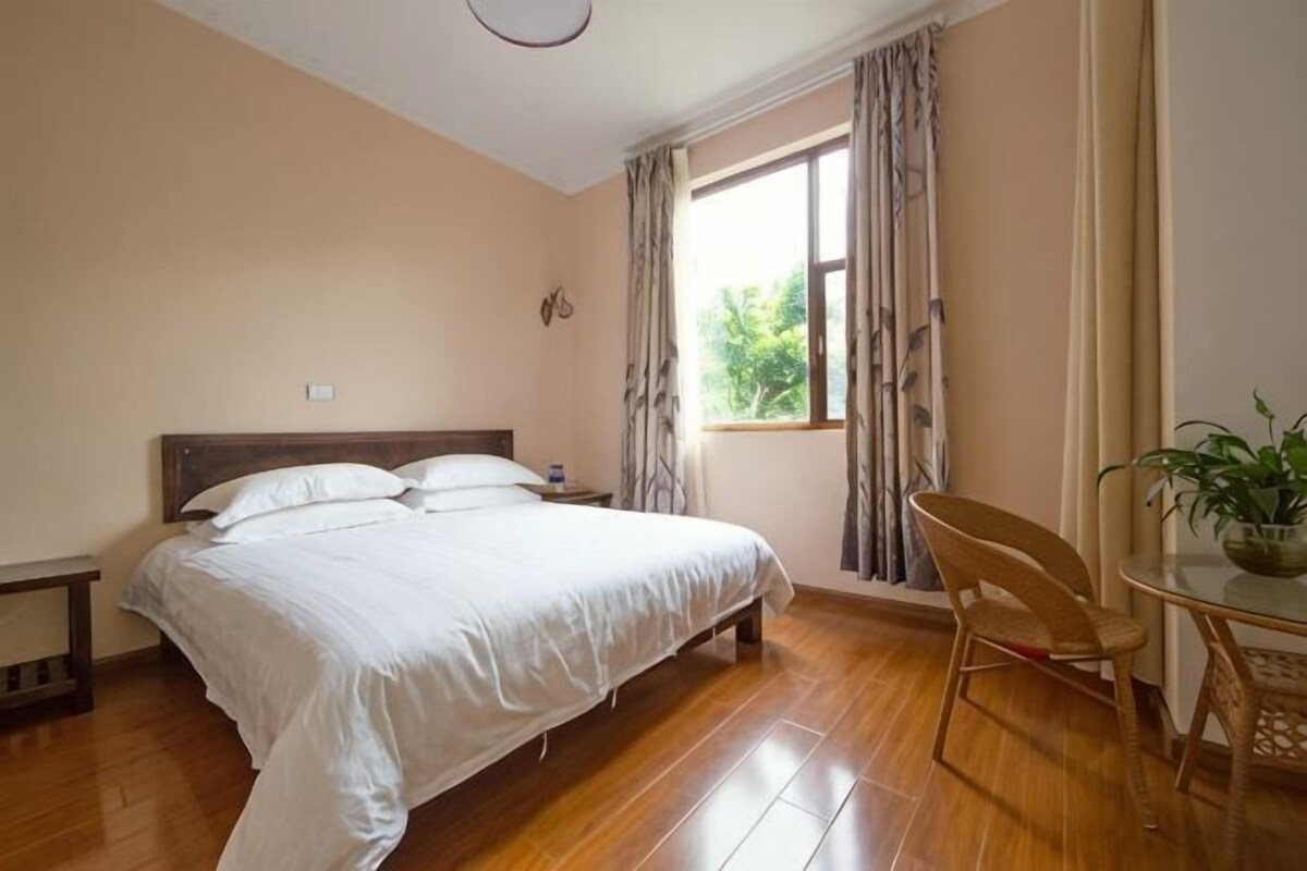 A bedroom with a bed, a chair, a table, a window with curtains, and a plant.