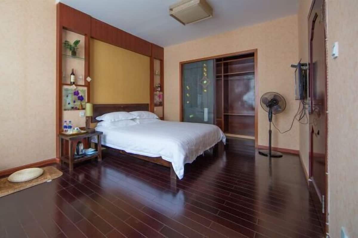 A hotel room with a bed, bedside table, wall-mounted shelf, and a standing fan.