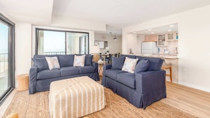 A modern living room with two dark sofas, a striped ottoman, and a large window. - Ocean Front Condo with Sunrise Views (Ocean City)