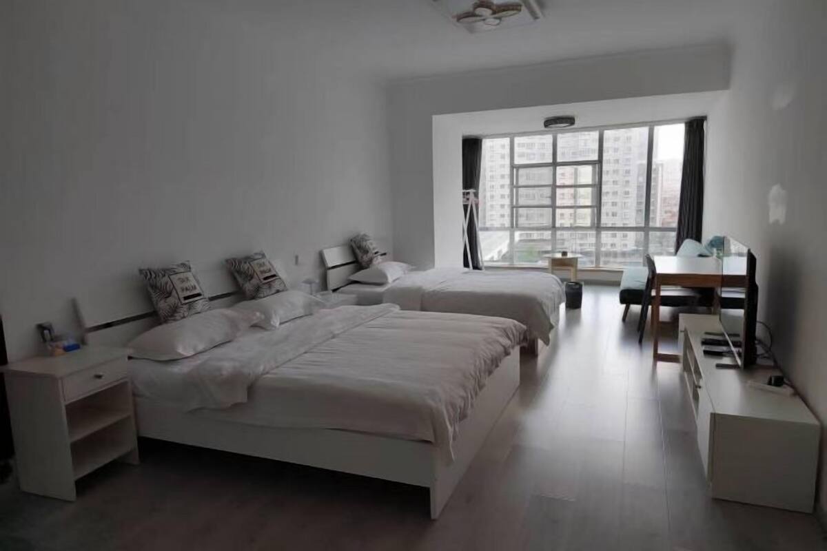 A modern bedroom with two beds, a desk, and a large window with a city view.