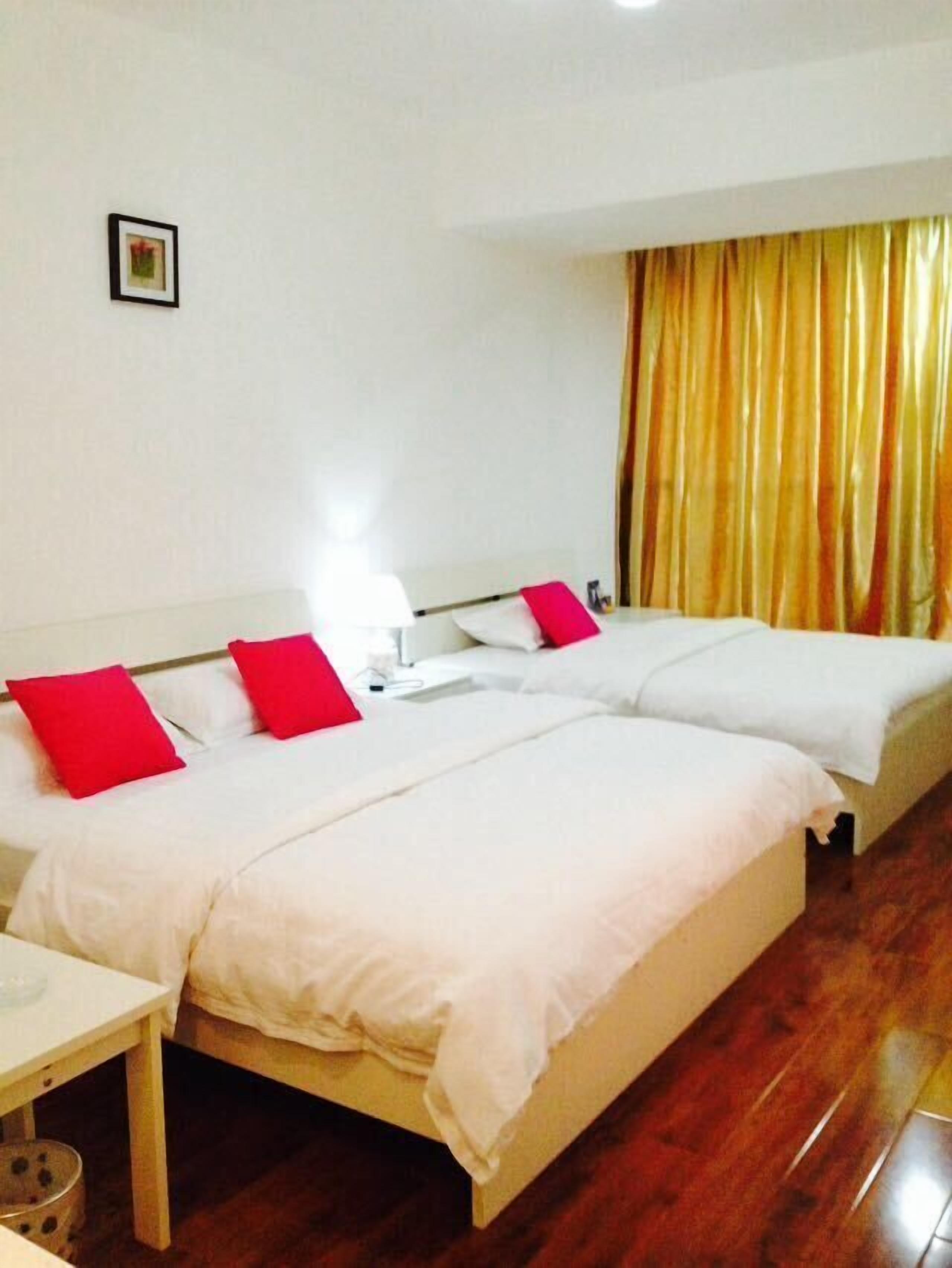 A hotel room with two beds, each with white bedding and red pillows, a small table, and a curtain with a golden hue.