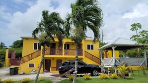 A two-story yellow building with a balcony, a car parked in front, and a hammock. - Cozy 1-bedroom apartment in peaceful Valley with AC, WiFi (Cassada Gardens)
