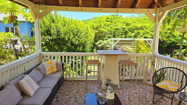 A covered outdoor seating area with a sofa, chairs, and a table, overlooking a lush garden and a blue building. - Cozy 1-bedroom apartment in peaceful Valley with AC, WiFi (Cassada Gardens)