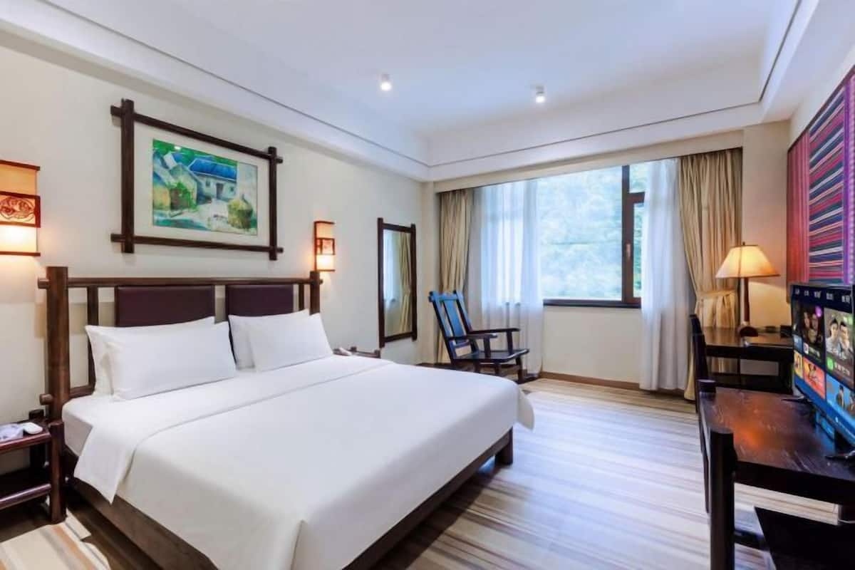 Ein Hotelzimmer mit einem großen Bett, einem Schreibtisch mit Computer, einem Stuhl und einem Fenster mit Vorhängen.